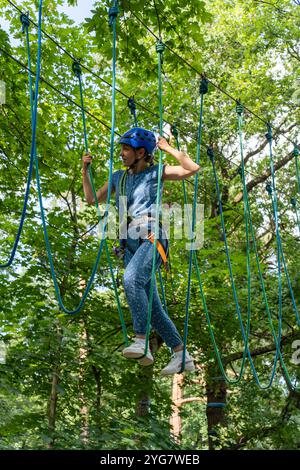 Donna sicura di sé con casco naviga su un percorso di corda alta nella vibrante foresta verde, bilanciandosi su una fune in un parco avventura all'aperto. Concetto di attivo Foto Stock