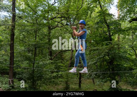 Donna sorridente in un percorso di corda con casco in alto sopra il suolo, in una foresta lussureggiante, in equilibrio su una corda con sicurezza. Avventura all'aria aperta, vita attiva Foto Stock