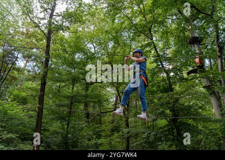 Donna con casco che naviga in corde in alto nella foresta, bilanciandosi su una corda con un'espressione mirata. Sport d'avventura, attività all'aperto e cha Foto Stock