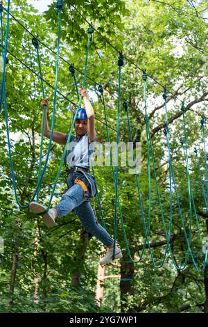 Donna sorridente in casco che si arrampica sulle corde in un parco avventura, in equilibrio su impegnativi percorsi a corde alte circondati da verde foresta nelle giornate di sole. Ciao Foto Stock