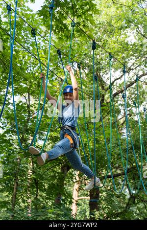 Donna felice con casco che naviga su un percorso di corda alta nella foresta verde, bilanciandosi con sicurezza sulle corde nel parco avventura all'aperto in una giornata luminosa e soleggiata. Fi Foto Stock