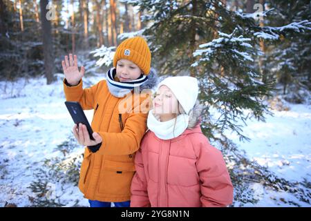 Un fratello e una sorella sono gioiosamente impegnati in una videochiamata, sullo sfondo mozzafiato di una foresta innevata. La scena cattura le loro risate e. Foto Stock