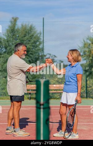 vertical prima della partita, un'anziana coppia di tennisti si salutano con un'amichevole stretta di mano, entrambi sorridenti. La rete dietro Foto Stock
