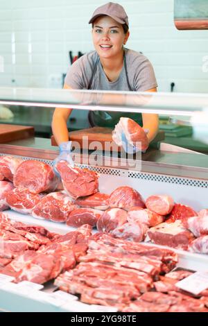 Il venditore giovane positivo in grembiule e guanti usa e getta mostra filetto di carne Foto Stock