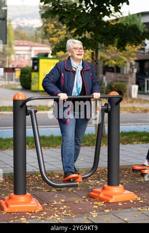 In un parco pieno di colori autunnali, una donna anziana con gli occhiali si esercita sulle attrezzature all'aperto. Il suo abbigliamento blu e la sua sciarpa completano la serena S Foto Stock