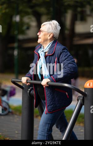 Una donna matura di settant'anni utilizza attrezzature da palestra all'aperto per mantenersi attivi in un tranquillo parco autunnale. Con un vestito casual blu e una sciarpa, lei Foto Stock