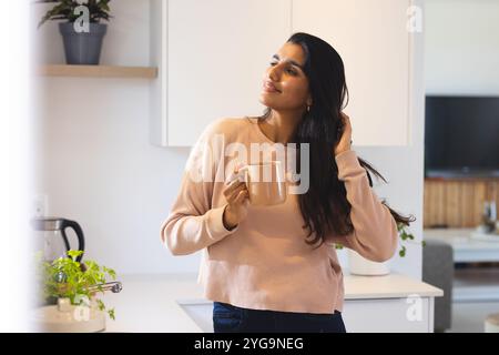 Donna asiatica che gusta il caffè mattutino in cucina, sorride e si sente rilassata, a casa Foto Stock