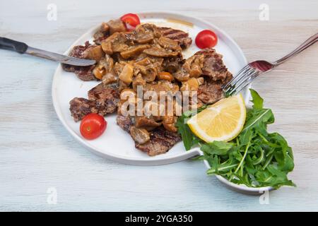 Bistecca alla griglia con salsa di funghi cremosa, servita con rucola, pomodori ciliegini e un spicchio di limone su un piatto bianco. Foto Stock