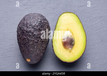 Tre avocado, uno intero e due metà, sono disposti su una superficie di ardesia grigia. Gli avocado tagliati a metà rivelano la loro cremosa carne gialla e la grande p marrone Foto Stock