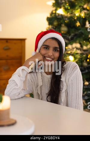 Donna sorridente nel cappello di Babbo Natale seduta accanto all'albero di Natale a casa Foto Stock