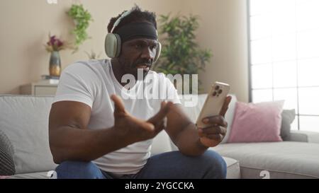 Un bel giovane afro-americano con la barba, con le cuffie, siede in salotto, regge uno smartphone, sembra frustrato o confuso Foto Stock