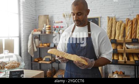 Bel giovane afroamericano che tiene una baguette in un'accogliente panetteria all'interno, con pane appena sfornato sugli scaffali sullo sfondo. Foto Stock