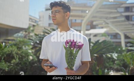 Giovane ispanico con in mano un mazzo di fiori e uno smartphone, in piedi in un parco urbano all'aperto con vegetazione, indossando una camicia bianca e occhiali, Foto Stock