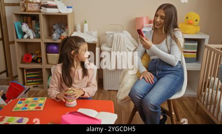 La mamma scatta una foto della figlia con il telefono al chiuso, catturando i momenti di amore della famiglia in un'accogliente stanza dei giochi piena di giocattoli e mobili. Foto Stock