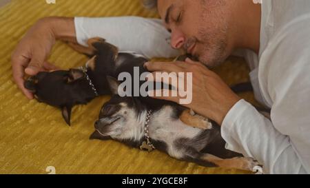 Uomo ispanico di mezza età che si rilassa a letto con due chihuahua a casa, mostrando un momento tenero con i suoi animali domestici in una camera da letto interna. Foto Stock