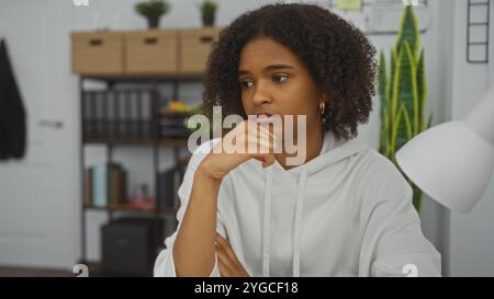 Giovane donna afro-americana con capelli ricci in un abbigliamento casual seduta con cura in un ufficio con sfondo da libreria. Foto Stock