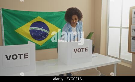 Una giovane donna afroamericana con i capelli ricci getta il suo voto in una sala universitaria elettorale brasiliana al coperto, con una bandiera brasiliana e una cabina di votazione pr Foto Stock