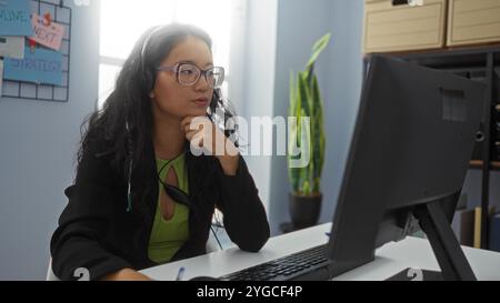 Donna che lavora in un ufficio con occhiali e cuffie, focalizzata sullo schermo del computer, seduta alla scrivania con attrezzature per impianti e ufficio, ur professionale Foto Stock