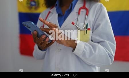 Una dottoressa afroamericana con un cappotto bianco usa il suo telefono al chiuso con una bandiera venezuelana sullo sfondo. Foto Stock