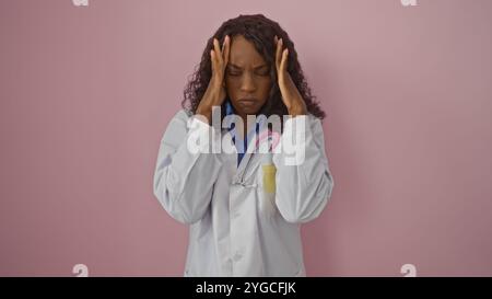 Una giovane dottoressa afroamericana frustrata con i capelli ricci in un cappotto bianco tiene la testa su uno sfondo rosa isolato. Foto Stock