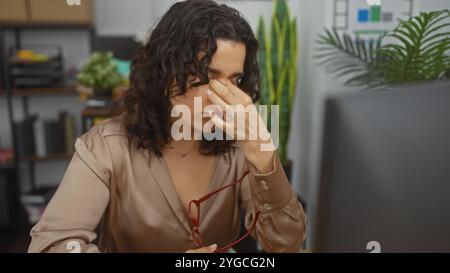 Una giovane e attraente donna ispanica in un ambiente d'ufficio, visibilmente stressata e con gli occhiali in mano, siede all'interno circondato da piante e luoghi di lavoro Foto Stock