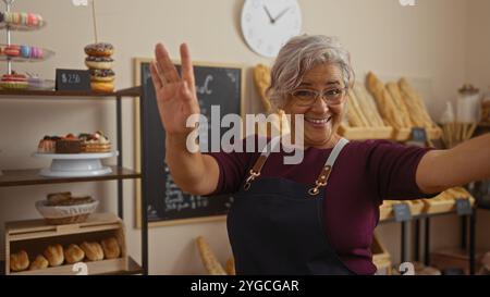 Donna ispanica che balla gioiosamente in una panetteria, circondata da pane e dolci, indossa occhiali e grembiule, trasuda felicità ed energia. Foto Stock