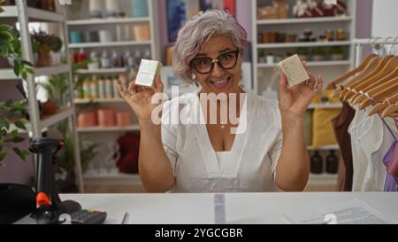 Donna anziana con capelli grigi e occhiali sorridenti mentre mostra saponette in un negozio moderno con scaffali e vestiti, isolato su sfondo rosa. Foto Stock