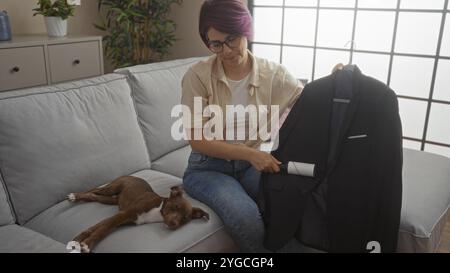 Una giovane donna caucasica si trova all'interno di un accogliente soggiorno, utilizzando un rullo di lanugine su una giacca nera accanto a un cane da letto sul divano. Foto Stock