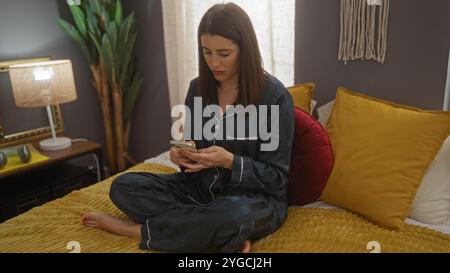 Una bella giovane donna ispanica in pigiama siede su un comodo letto in una camera da letto elegante, utilizzando con attenzione il suo smartphone nella calda luce di una sera Foto Stock
