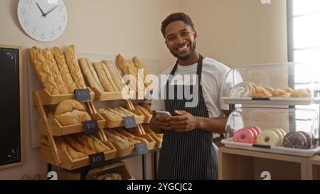 Giovane sorridente in panetteria con pane sullo sfondo e pasticcini in esposizione mentre tiene in mano un telefono Foto Stock