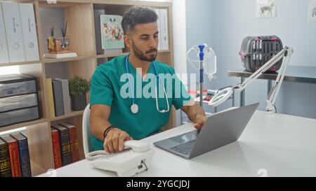 Un bel giovane veterinario ispanico maschile in scrub verdi lavora su un computer portatile e risponde a un telefono in una clinica veterinaria luminosa e organizzata. Foto Stock