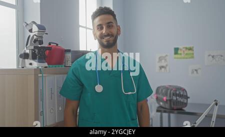 Un bellissimo giovane veterinario ispanico in scrub verdi con uno stetoscopio intorno al collo sorride in una luminosa stanza veterinaria con vari tipi di animali Foto Stock