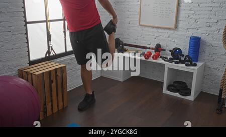 Un uomo allunga la gamba in una palestra interna ben attrezzata con vari pesi e attrezzi per il fitness esposti su scaffali contro un muro di mattoni. Foto Stock