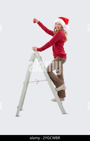 Buon Natale, felice donna sorridente decora l'albero di Natale, sale su una scala di legno vintage con la palla di Natale in mano, guardando la cameriera Foto Stock