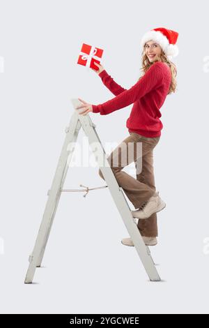 Buon Natale, felice donna sorridente decora l'albero di Natale, sale su una scala in legno vintage con una confezione regalo rossa in mano, guardando la macchina fotografica Foto Stock