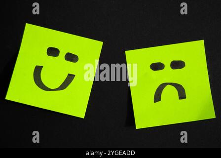 Una foto concettuale che mostra due note adesive verdi: Una con il volto sorridente e l'altra con una triste emoji, su uno sfondo neutro. Foto Stock