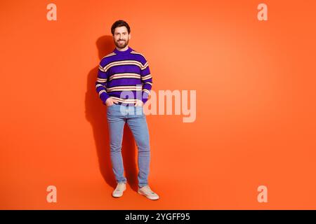 Il giovane uomo barbuto in un elegante pullover a righe si erge con sicurezza su un vivace sfondo arancione, simboleggiando la moda moderna e l'energia giovanile. Foto Stock