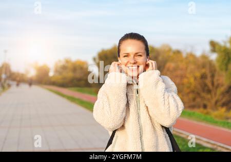 Gioiosa giovane donna bianca in giacca e auricolari beige che guarda la macchina fotografica e sorride mentre cammina all'aperto nel parco. Foto Stock