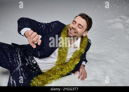 Un giovane gioioso ride mentre sdraiato sulla neve, adornato da allegre decorazioni natalizie intorno a lui. Foto Stock