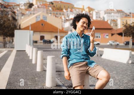 Un giovane con i capelli ricci neri, con una camicia in denim e pantaloncini marroni, siede all'aperto su un palo di cemento, sorridendo al suo smartphone; ambientazione urbana Foto Stock