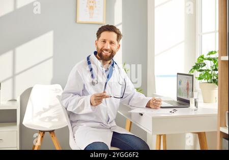 Seduto al tavolo, il medico guarda la macchina fotografica con un sorriso felice. Uomo medico impegnato in medicina trasuda professionalità e compassione nel fornire assistenza Foto Stock