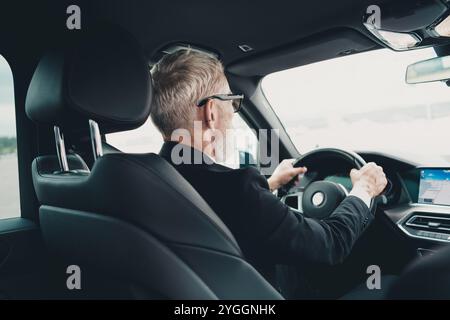 Uomo d'affari maturo che guida in città, concentrato sui viaggi. Tuta professionale al volante di un'auto di lusso, che incarna il successo. Foto Stock