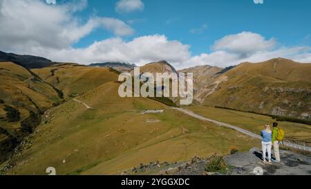 Due viaggiatori ammirano la vista panoramica mozzafiato delle colline verdi ondulate e delle aspre cime montuose che circondano la chiesa della Trinità di Gergeti a Kazbeg Foto Stock