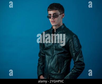 Giovane uomo elegante con occhiali da sole e giacca in pelle che posa con sicurezza su uno sfondo blu vivace Foto Stock
