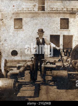 Questa è una foto originale e reale di un uomo in abito del XIX secolo, tra cui un cappello da bocciatore, che si trova accanto a grandi ingranaggi e tubi su quello che sembra essere il ponte di una nave a vapore. Il contesto marittimo e i suoi vestiti suggeriscono un periodo di tempo tra la fine degli anni '1800 e l'inizio degli anni '1900, probabilmente durante il picco delle navi alimentate a vapore. L'immagine offre uno scorcio raro sulla storica tecnologia marittima e sulla vita delle navi, preservando un pezzo della storia industriale e marittima dell'epoca. Foto Stock