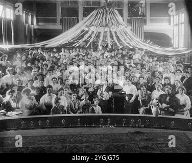 Questa è una foto originale e reale di un grande raduno di donne nell'America dei primi anni del XX secolo, probabilmente dell'epoca edoardiana (circa 1900-1915). La sala è decorata con bandiere e rastrelli americani, suggerendo un evento patriottico o formale. L'abbigliamento femminile riflette lo stile del periodo, catturando un momento di impegno sociale e comunitario nella prima società americana. Foto Stock