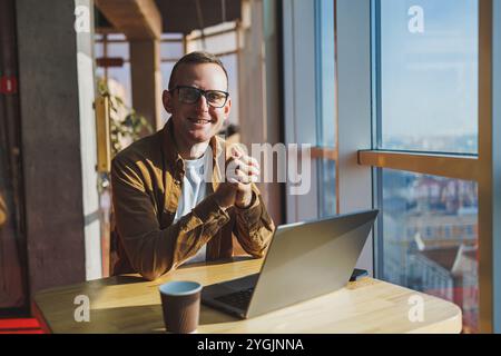 Un uomo equilibrato con occhiali e abiti casual lavora su un laptop da un'area di lavoro accogliente. Un freelance di successo lavora in remoto. Foto Stock