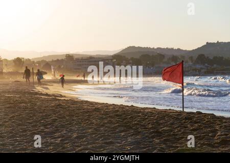 Persone irriconoscibili che camminano su una spiaggia con una bandiera rossa che avverte delle grandi onde all'alba Foto Stock