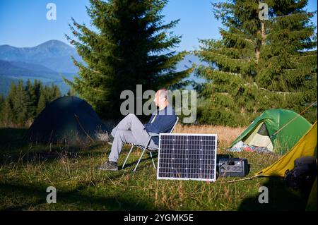 Uomo che utilizza un pannello solare fotovoltaico per caricare una centrale elettrica portatile all'aperto in campeggio ecologico. Il turista maschile siede rilassato su una sedia vicino alle tende. Sullo sfondo alti pini e cielo azzurro. Foto Stock