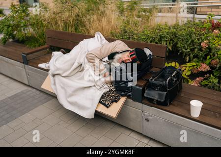 Senzatetto anziano esausto che dorme tranquillamente su una panchina intemprata fuori nelle strade urbane della città durante la giornata fredda. Uomo mendicante stanco e anziano che giace all'aperto. Foto Stock